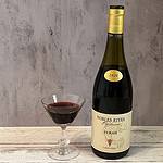 Bottle of Nobles Rives 2020 Syrah. Red wine ina crystal wine glass alongside wine bottle with gold foil capsule around the neck and beige lablel.