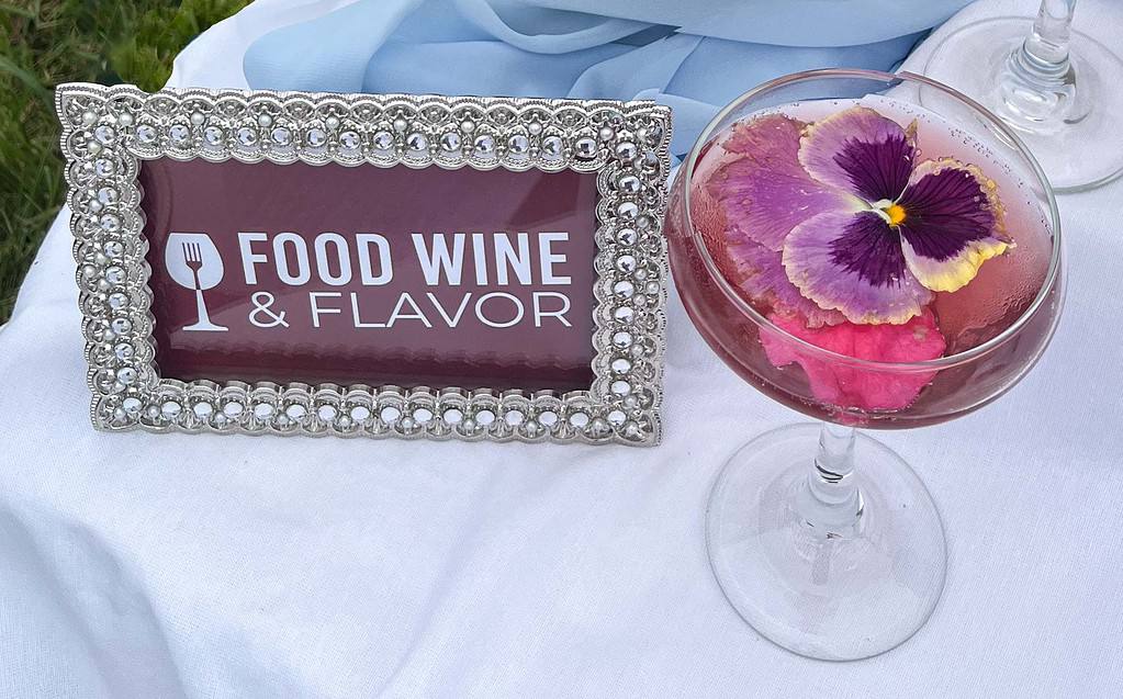 Food WIne and Flavor logo on a maroon background inside a clear stone embellished frame. To its right is a couple glass filled with a cranberry-colored liquid. Floating in the glass is a rose petal anda pansy.