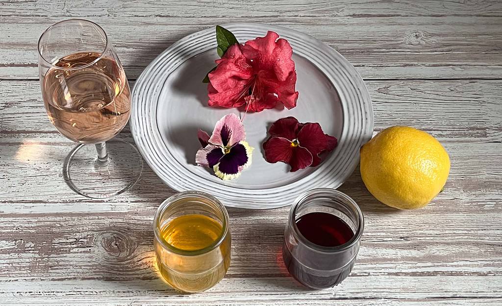 White plate with pansy, pink flower and varied pansy. Rose wine in wine glass, lemondrop mix, pomegranate cocktail mix and a whole lemon