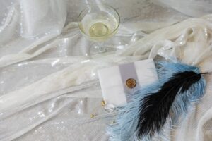 White silky fabric. On top center is coupe glass of Champagne, Center right is a white envelope with a narrow gray paper strip sow the middle, sealed with gold seal. Bottom right are two feathers - one sky blue and one black.