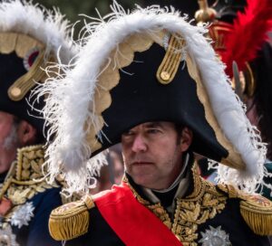 Close-up of an actor dressed as Napolean Bonaparte whereing a tradtional French military uniform with navy blue, gold espelettes and characteristic bell-shaped hat lined with white fur.