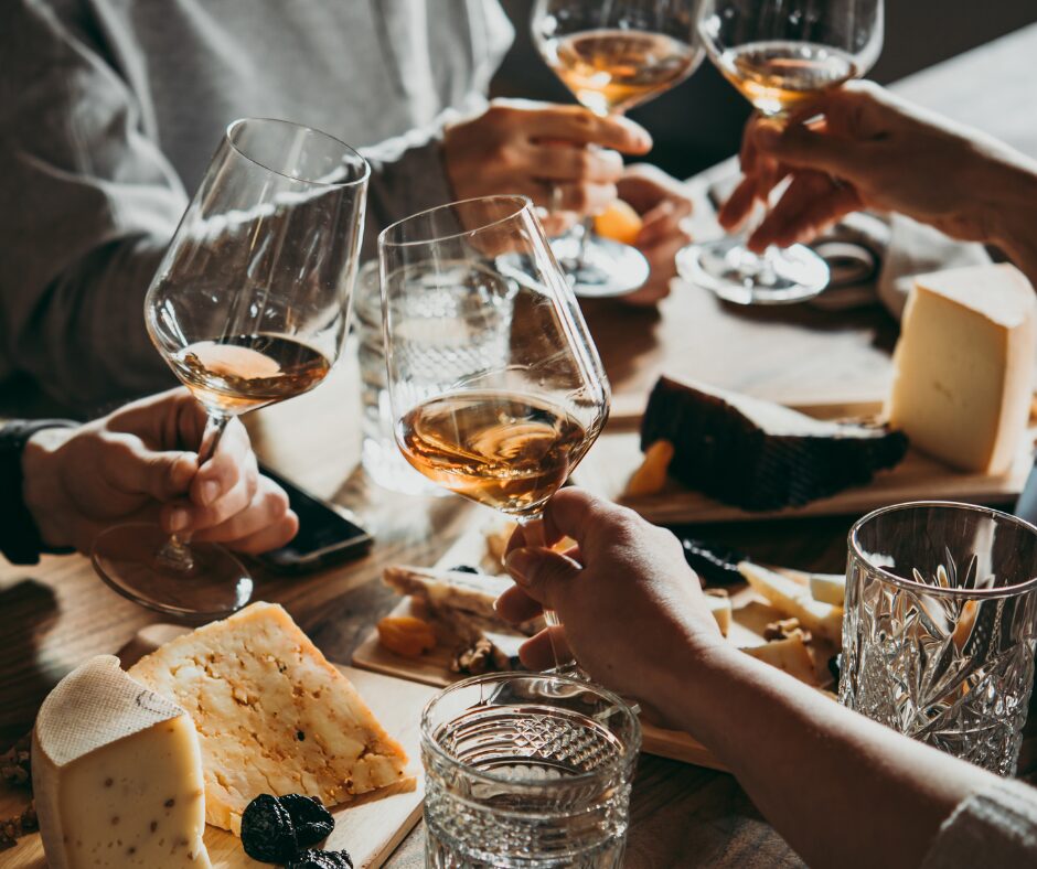 four friends cliinking wine glasses over a table or cheeses. Non of the people can be seen beyond their hands.