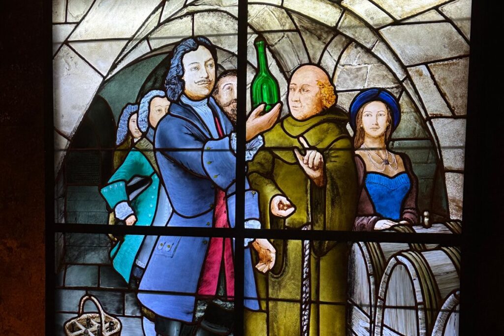 Stained glass window with a wealthy landowner holding up a wine bottle in front of a group of citizens and a Benedictine monk.