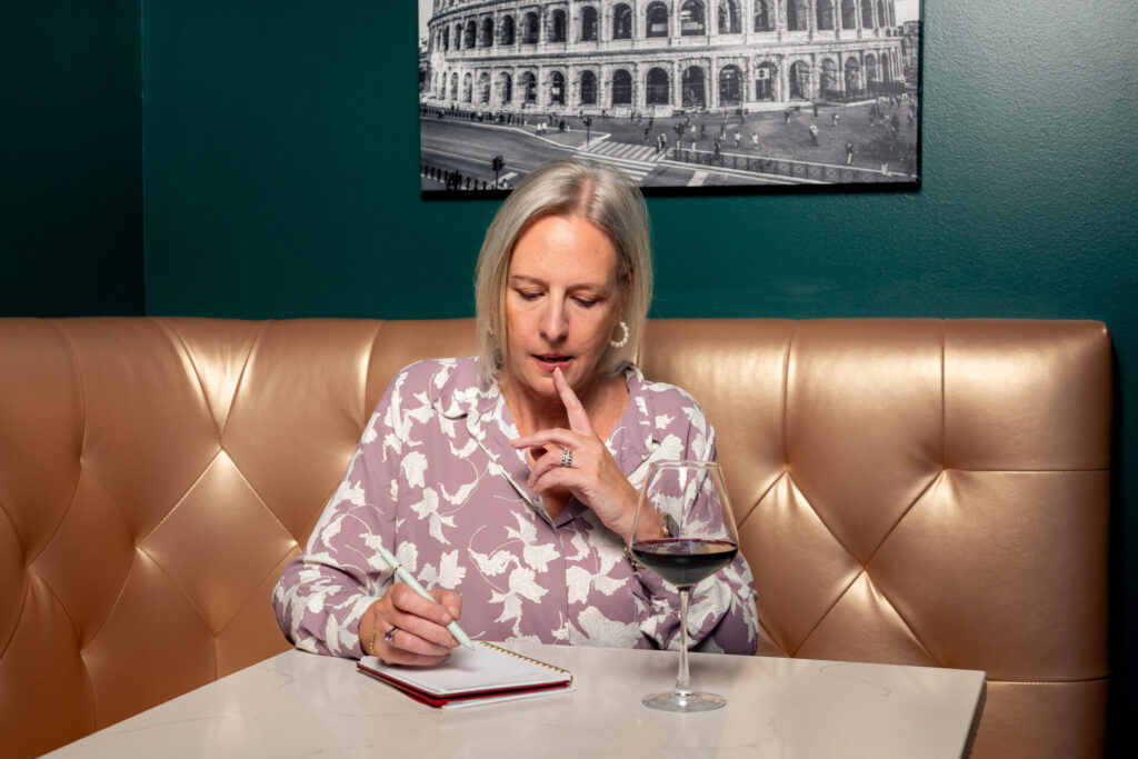 Anne Sitting in a large booth, pointer finger on her lower lip as she gazes down at her notebook, thinking about what to write.