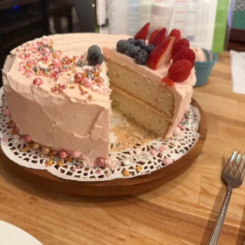 Two-tiered White Cake frosted with blush pink frosting and topped with fresh berries. Decorated with pink and gold cake decor.
