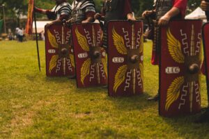 Line of four Roman soldiers. Photo frames the shields at their feet. We do not see their heads nor faces.