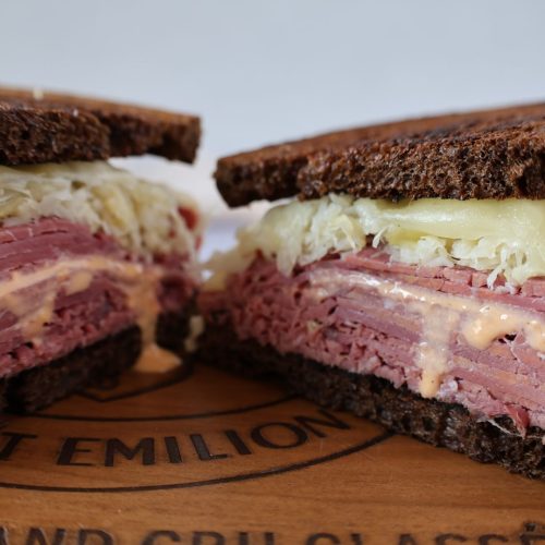 photograph of a Rueben sandwich cut in half and open-faced. Dark rye bread and piles of sliced corned beef, sauerkraut, swiss cheese and Russian dressing dripping down to the wood board.