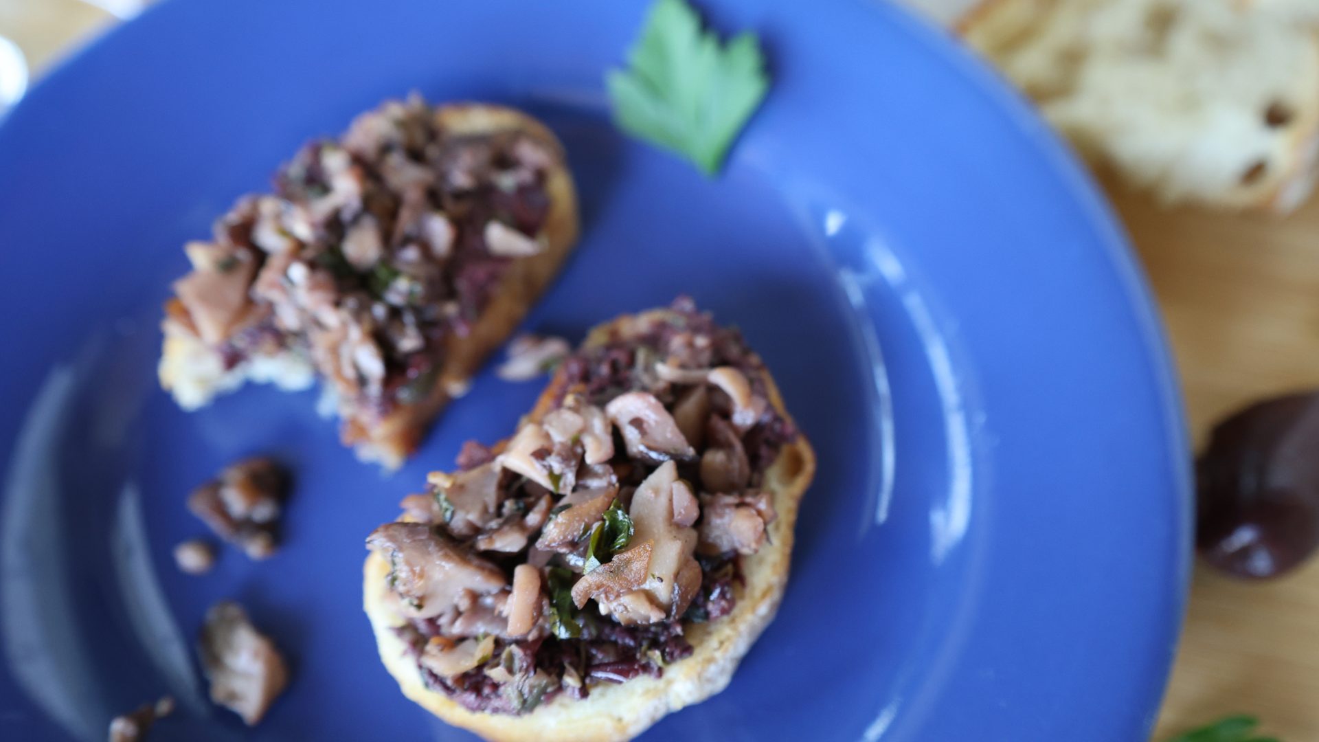 Royal blue plate with two crostinis of mushroom and olive tapenade with a sprig of parsley on the plate