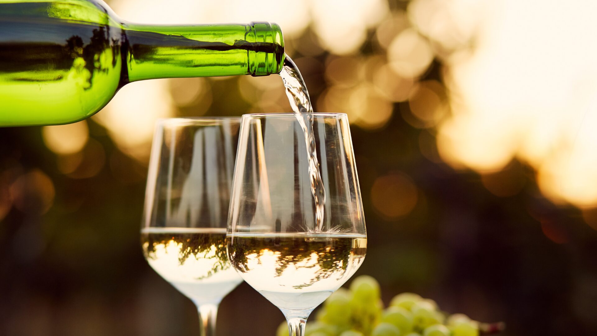 White Wine Glass Pouring 1920×1080 Green glass wine bottle pouring golden Riesling into two wine glasses side by side, filling halfway to the widest part of the glass. Backlit by the sun.